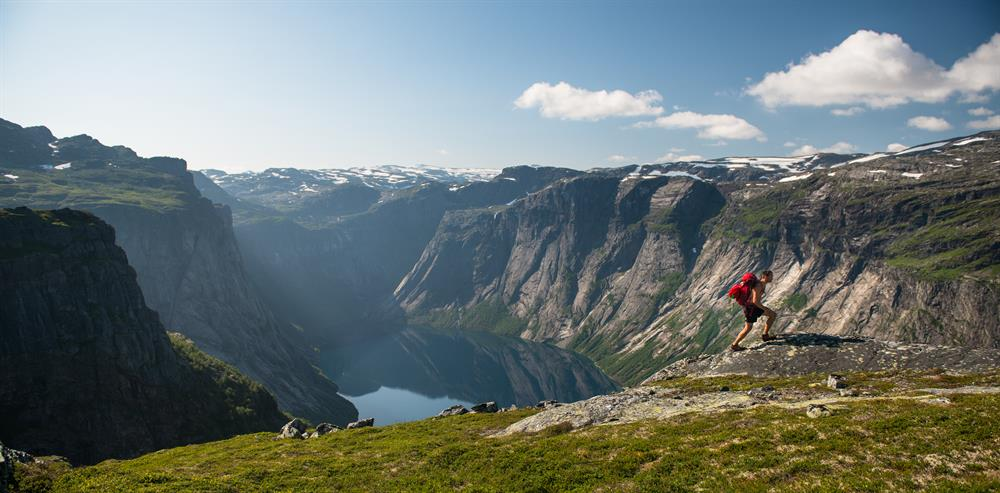 Trekker di chuyển trên cung đường Trolltunga vào mùa hè, trời trong xanh, cảnh quan núi non và tuyết tan xen lẫn thảm cỏ
